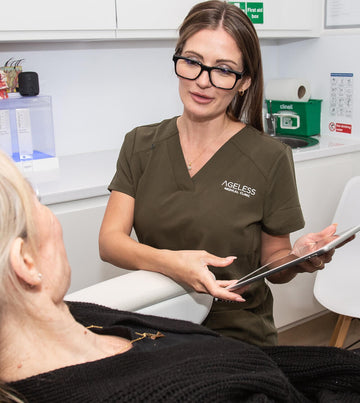 Woman in a brown 'Ageless Medical Clinic' scrub top holding a tablet in a clinical setting.